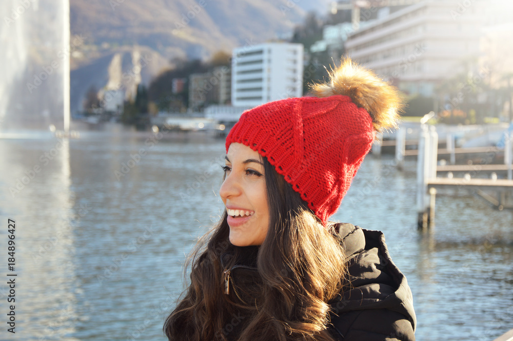 Happy winter woman with red wool hat and pompon with the city lake on the background - obrazy, fototapety, plakaty 