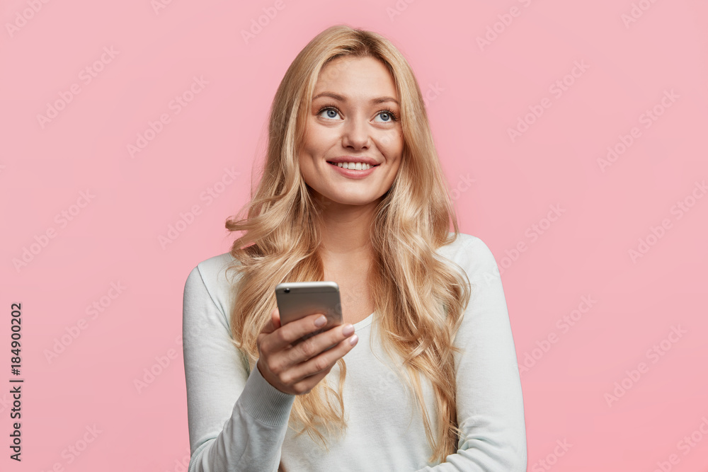 Cute dreamful smiling young beautiful female holds modern smart phone reads news on website, has joy as enjoys online communication with friends via social networks, isolated over pink background. Cute dreamful smiling young beautiful female holds modern smart phone reads news on website, has joy as enjoys online communication with friends via social networks, isolated over pink background.