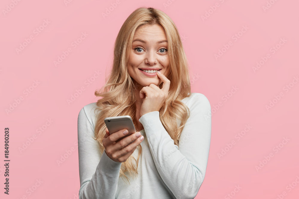 Smiling blonde female blogger uses smart phone for surfing her webpage, being satisfied with website popularity, has many feedbacks from followers, connected to wireless internet, isolated on pink Smiling blonde female blogger uses smart phone for surfing her webpage, being satisfied with website popularity, has many feedbacks from followers, connected to wireless internet, isolated on pink