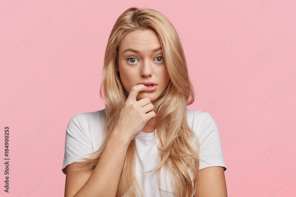 Beautiful female with puzzled expression, keeps finger on lips, listens interlocutor in bewilderment, poses against pink background. Studio shot of frustrated blonde woman with pure healthy skin Beautiful female with puzzled expression, keeps finger on lips, listens interlocutor in bewilderment, poses against pink background. Studio shot of frustrated blonde woman with pure healthy skin