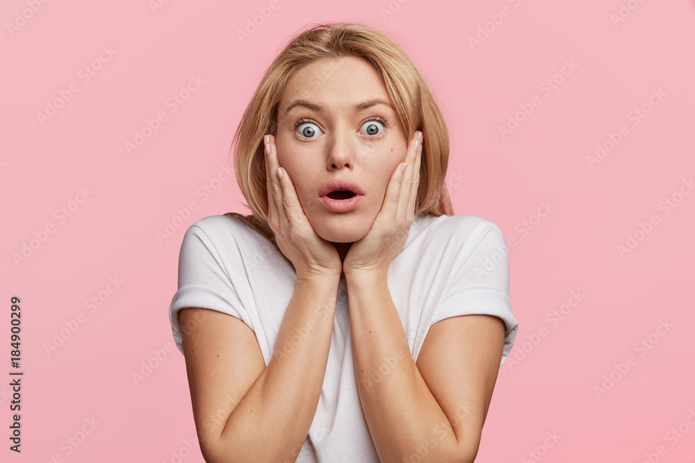 Scared European woman looks with blue bugged eyes at camera, being afraid of something, keeps hands on cheeks, looks in terror, isolated over pink background. Emotional horrified female indoor Scared European woman looks with blue bugged eyes at camera, being afraid of something, keeps hands on cheeks, looks in terror, isolated over pink background. Emotional horrified female indoor