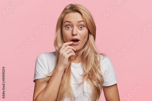 Surprised positive female with blue eyes popped out, opens mouth widely in surprisment as hears news from interlocutor, straes at camera, expresses shock and disbelief, isolated on pink wall