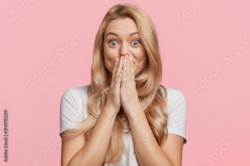 Beautiful excited blonde female covers eyes with happiness, doesn`t expect to recieve pleasant surprise from close friend, being thankful and pleasantly shocked, isolated over pink background photo