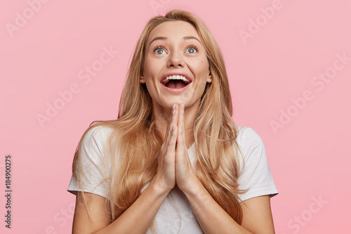Positive hopeful female with blonde long hair, keeps palms together, being in high spirit, looks with excitement upwards, isolated over pink background. Please, help me. Woman begs in studio
