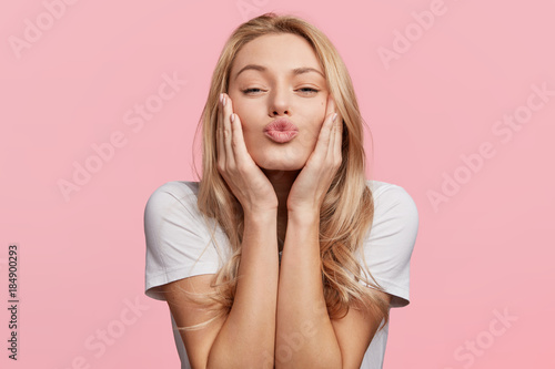 Portrait of lovely blonde female rounds lips as going to recieve kiss from boyfriend, looks at camera with pleased expression, isolated over pink background. Beautiful young woman poses in studio
