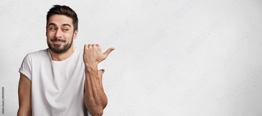 Horizontal portrait of cheerful hesitant male with thick beard and mustache wears casual t shirt, indicates with thumb aside, stands against white concrete background, copy space for advertising - obrazy, fototapety, plakaty 