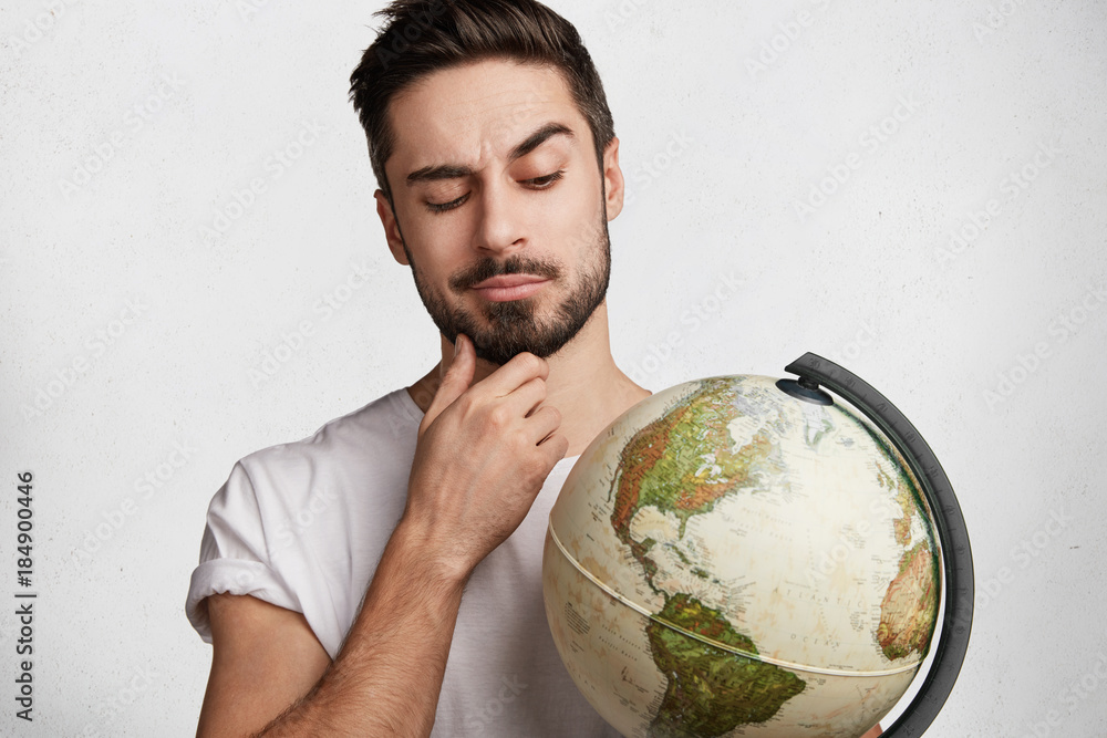 Portrait of thoughtful bearded businessman wants to explore unknown places,  looks at worldwide globe, tries to choose destination, isolated over white  concrete wall. Young male holds earth indoor Stock Photo | Adobe