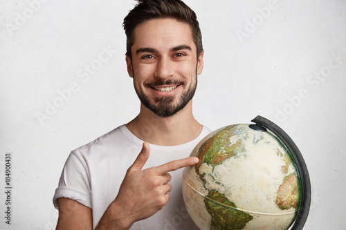 Handsome cheerful young active bearded traveller holds globe, chooses destination where to go, indicates at different continents, makes important choice. People, travelling and adventure concept photo