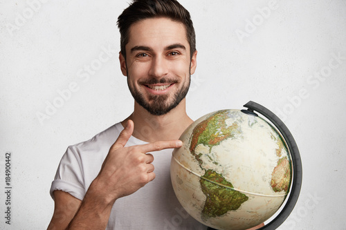 I choose this place. Glad positive male with beard and mustache wears casual t shirt, points at destination on globe, being happpy to win tourist voucher in exotic country. Travelling concept photo