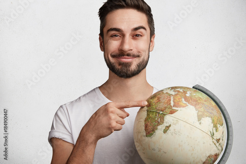 Shot of positive man points at Africa on globe, being glad to have vacations and opportunity to spend free time there, isolated over white concrete wall. Male student with orb. Geography concept photo