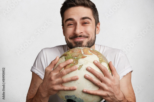 Pleased glad male with happy expression, embraces globe as rejoices holidays, wants to travel somewhere. Positive unshaven handsome man expresses love to universe. I love all world which surround me photo