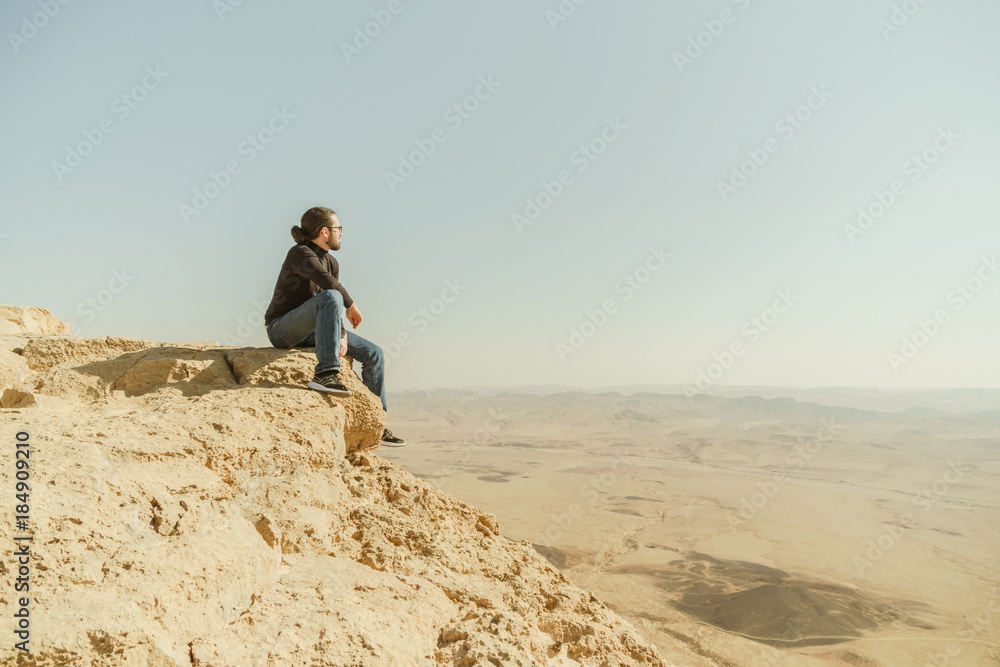 Extreme young man sitting on the edge of cliff and looking on desert