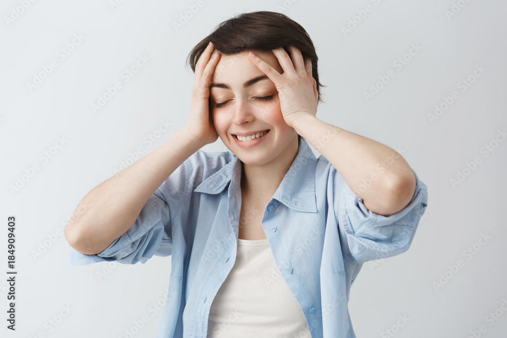 Joyful young student girl with dark short hair and closed eyes, holding head with hands, being super excited about birthday present from boyfriend. Joyful young student girl with dark short hair and closed eyes, holding head with hands, being super excited about birthday present from boyfriend.