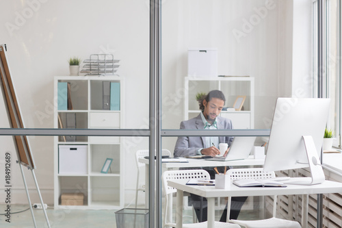 Young manager working behind transparent wall with empty workplace of another specialist near by