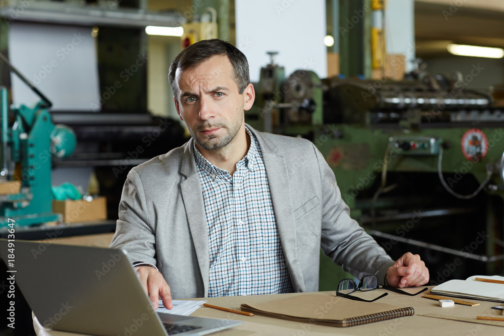 Serious foreman sitting by table in front of laptop and looking at camera while networking Serious foreman sitting by table in front of laptop and looking at camera while networking