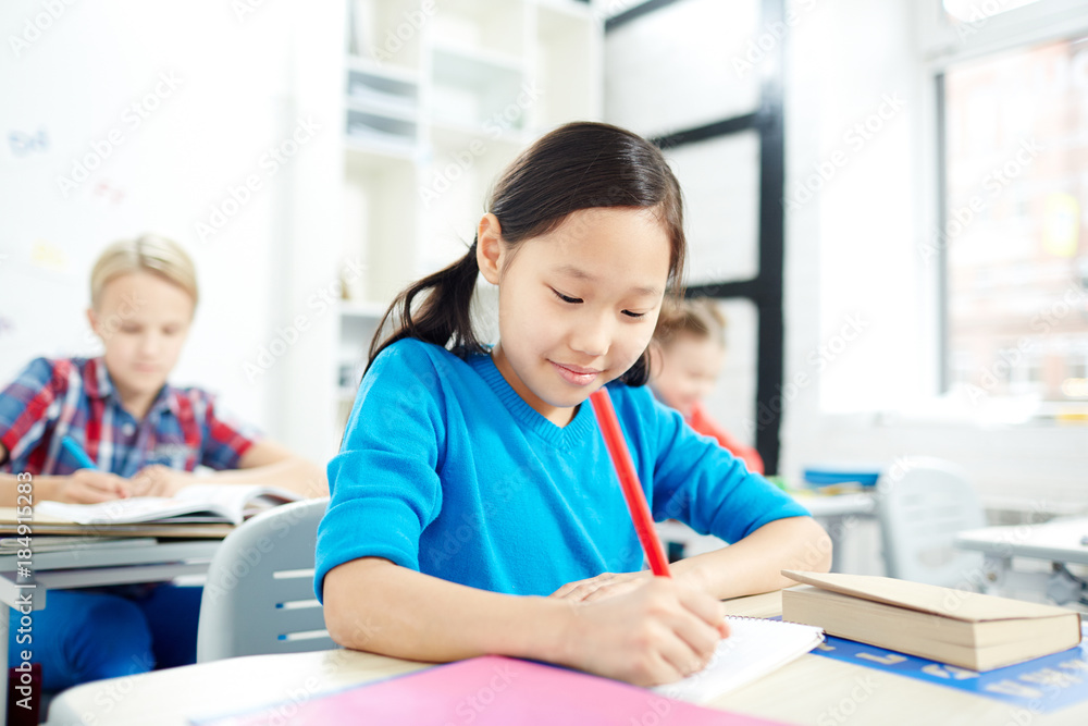 Little Asian primary school girl with red crayon drawing by desk at lesson Little Asian primary school girl with red crayon drawing by desk at lesson