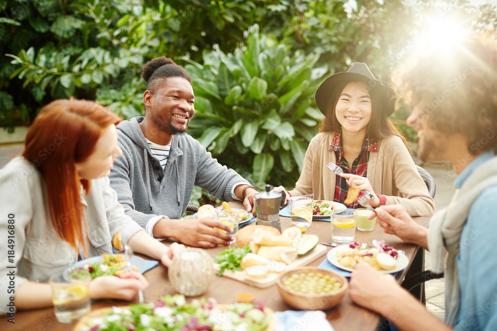 Group of young cheerful friends gathered by dinner table having talk ahile eating homemade food