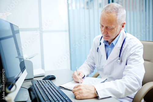Mature practitioner making notes or prescriptions in medical form by his workplace