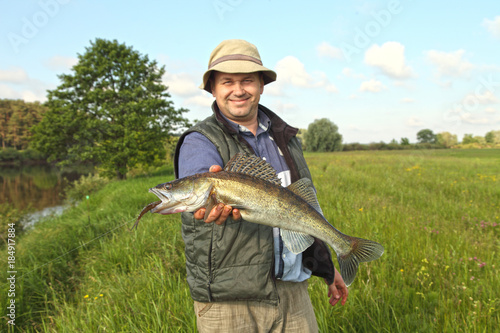 The fisherman holds a pike perch in the hands against the background of the summer landscape. Fishing is a man's hobby.