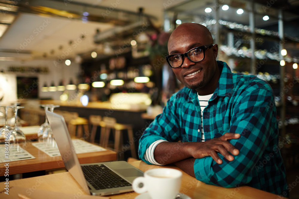 Confident businessman in eyeglasses sitting in front of laptop in cafe and looking at camera Confident businessman in eyeglasses sitting in front of laptop in cafe and looking at camera