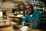 Confident businessman in eyeglasses sitting in front of laptop in cafe and looking at camera