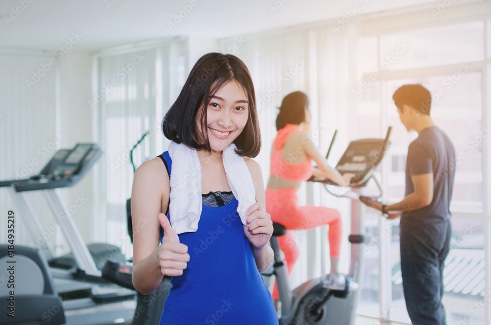 young happy asian woman slim body with towel showing thumbs up in fitness gym with people on machine bicycle background, bodybuilder, healthy lifestyle, exercise, workout and sport training concept young happy asian woman slim body with towel showing thumbs up in fitness gym with people on machine bicycle background, bodybuilder, healthy lifestyle, exercise, workout and sport training concept