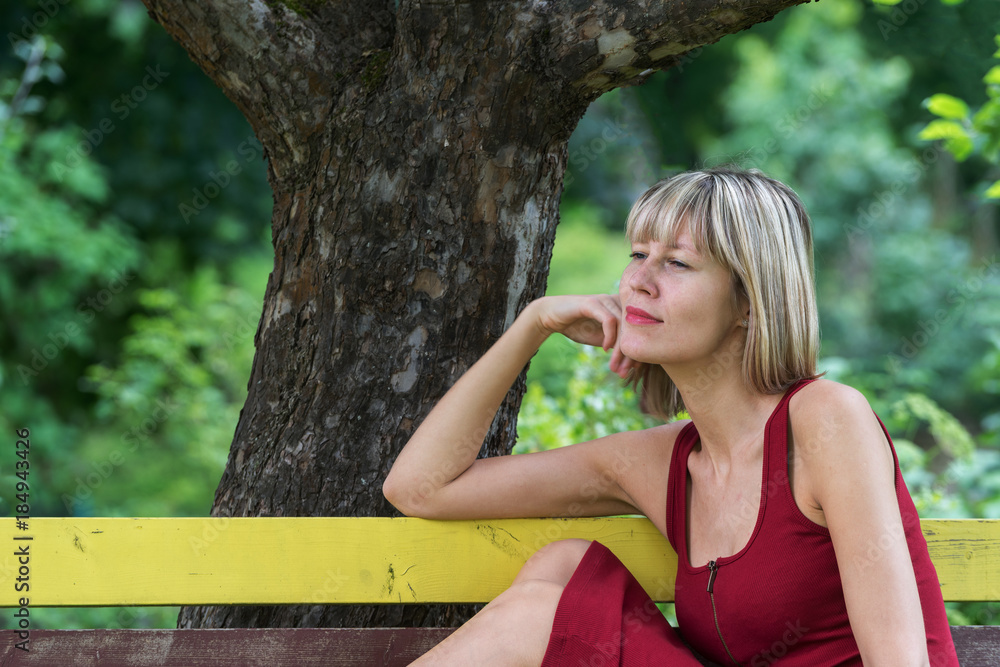 Naklejka premium Young blond woman in a red dress leaning sits on a wooden bench. .Businesswoman walking along the street and resting in the green city park on the beautiful sunny summer day.
