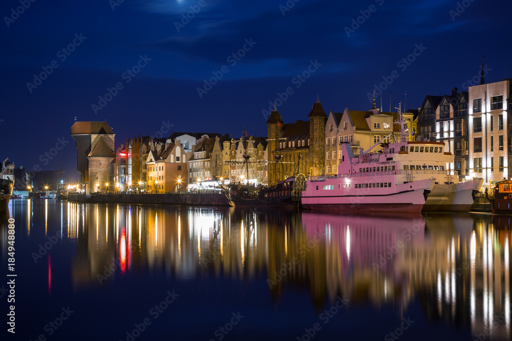 View of two boats and lit old buildings at the Long Bridge waterfront at the Main Town (Old Town) in Gdansk, Poland, in the evening.