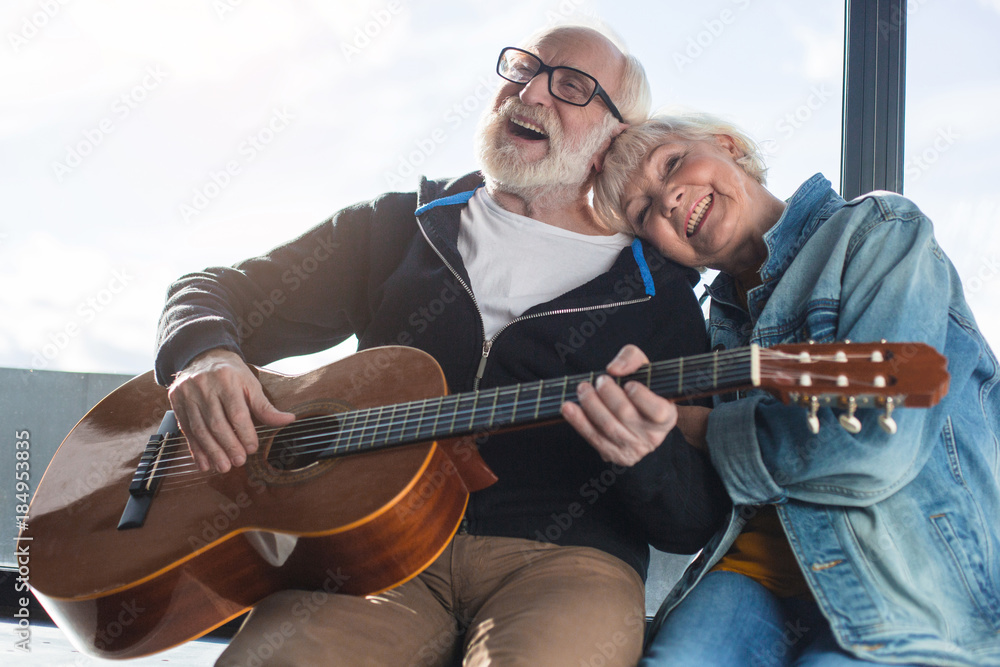 Romantic moments. Waist up portrait of two mature pensioners sitting on windowsill. Man is performing composition on guitar