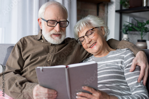 Waist up portrait of old married man and woman smiling with closed eyes. Husband is hugging wife while sitting at living room. Woman is holding photo album