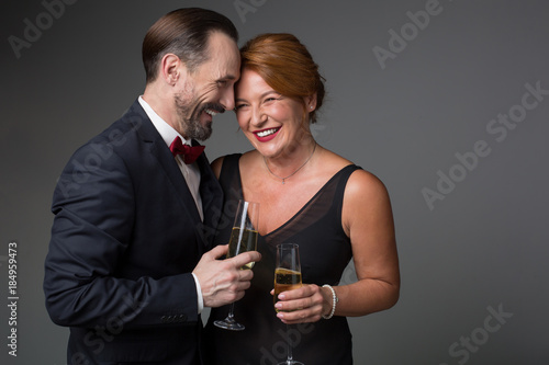 Portrait of cheerful middle-aged lovers are embracing and laughing. They are standing in elegant clothing and holding glasses of champagne. Isolated and copy space