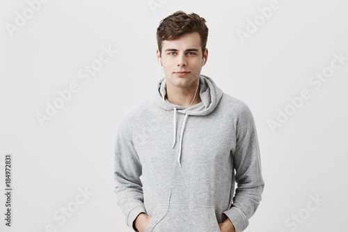 Handsome attractive European man in gray hoodie, with hands in pockets, look pleased at camera, has good mood as comes home after work. Good-looking male student poses against gray studio wall.