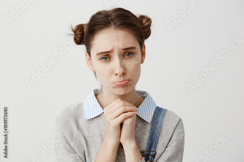 Close up portrait woman with brown hair in double buns pouting with pity look holding hands like praying. Pathetic expressions of girl asking for forgiveness over white wall. Concept of emotions