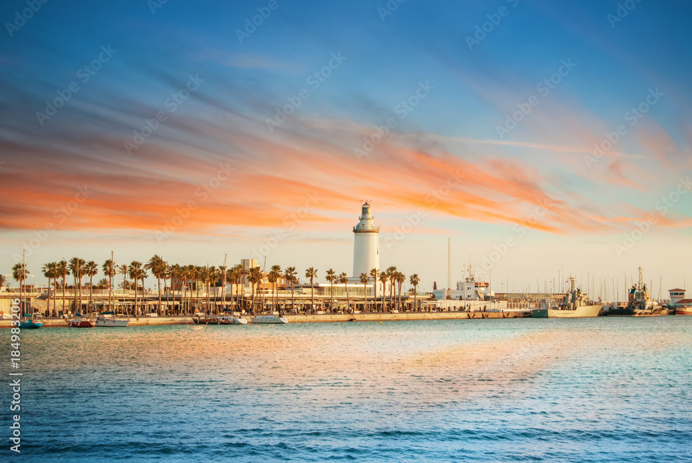 Evening panoramic aerial top view to a promenade with palms, shops and yachts and a lighthouse by the sea with dramatic colorful evening sunset sky at the background. Holiday vacation by the sea.