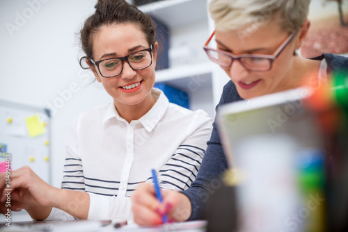 Fotografie Close up of two charming satisfied professional women sitting in the office and working together one next to another