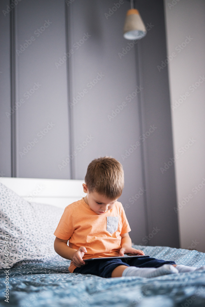 Playful cute curious little toddler boy sitting on the bed with a tablet in hands and playing video games alone. Playful cute curious little toddler boy sitting on the bed with a tablet in hands and playing video games alone.