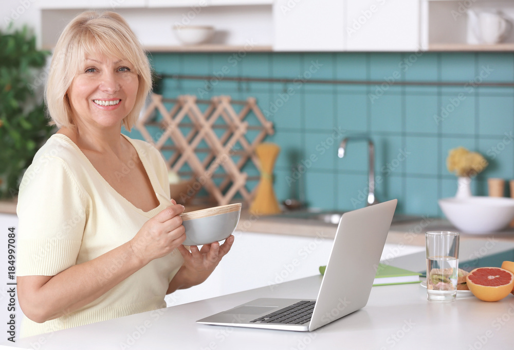 Mature woman having breakfast while working on laptop in kitchen Mature woman having breakfast while working on laptop in kitchen