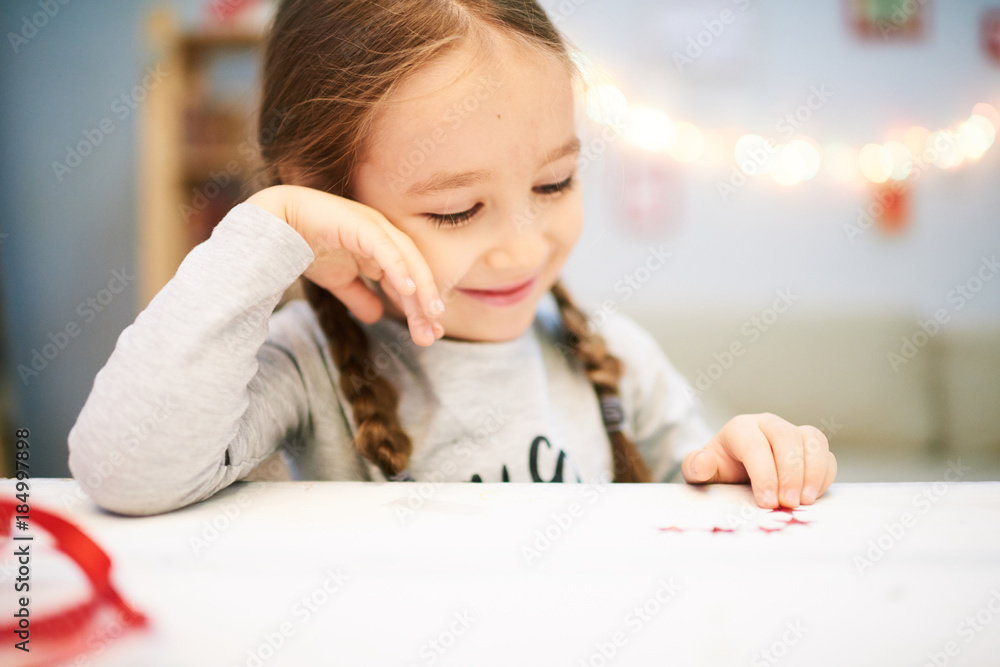 Head and shoulders portrait of adorable little girl with two braids sitting at desk in cozy bedroom and enjoying beauty of just finished Christmas card Head and shoulders portrait of adorable little girl with two braids sitting at desk in cozy bedroom and enjoying beauty of just finished Christmas card