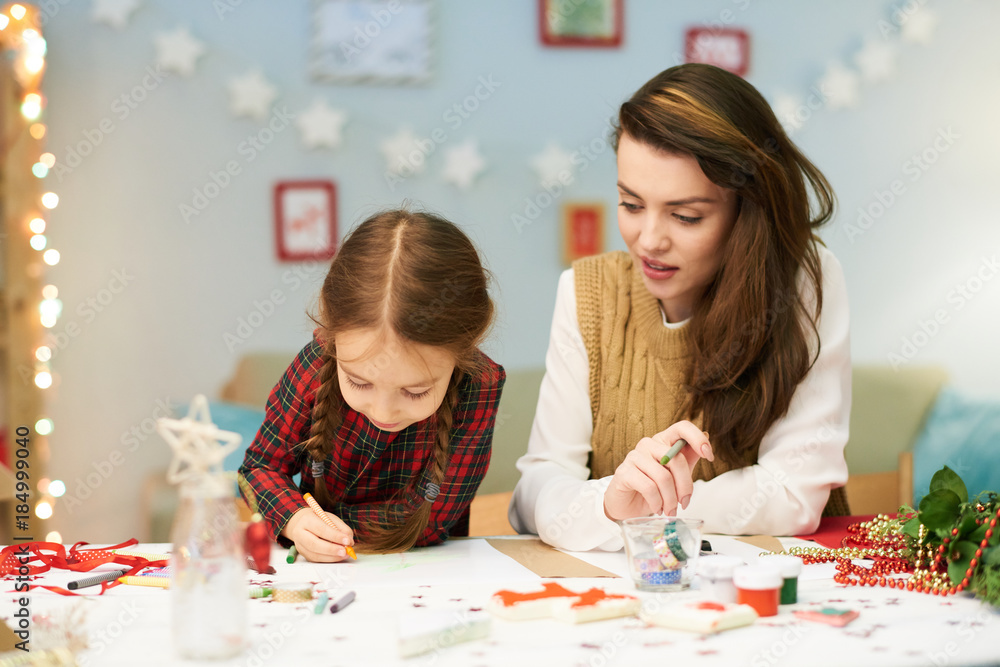 Fototapeta premium Little girl wrapped up in drawing picture with wax crayons while sitting at desk, her beautiful mother keeping eye on her, interior of living room decorated for Christmas celebration on background