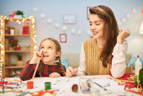 Dreamy little girl distracted from drawing colorful picture and looking upwards with interest  her attractive mother sitting next to her at desk  interior of decorated living room on background