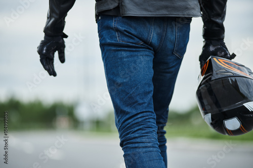 Cropped outdoor portrait of brutal young European man going in black leather jacket and wit halmet in the hand. Concept of hard and extreme hobby. Back view on motorbiker. photo