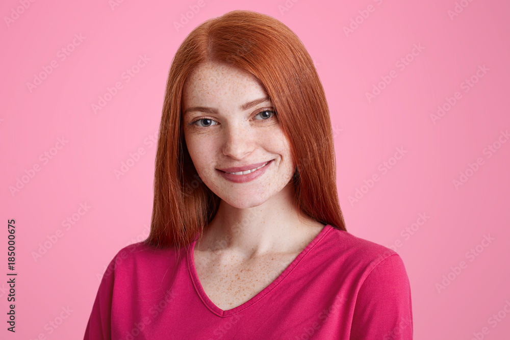 Close up portrait of smiling freckled female with straight luxurious red hair, being glad to recieve good job proposal, rejoices her success, isolated over pink background. Facial expressions concept Close up portrait of smiling freckled female with straight luxurious red hair, being glad to recieve good job proposal, rejoices her success, isolated over pink background. Facial expressions concept