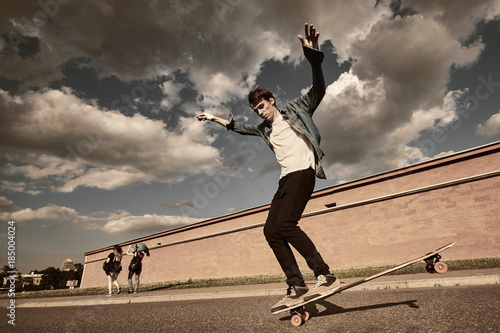 Lifestyle shot of fashionable young male in sneakers, black skinny jeans, white t-shirt and denim shirt enjoying free riding on skateboard in city surroundings against cloudy sky background