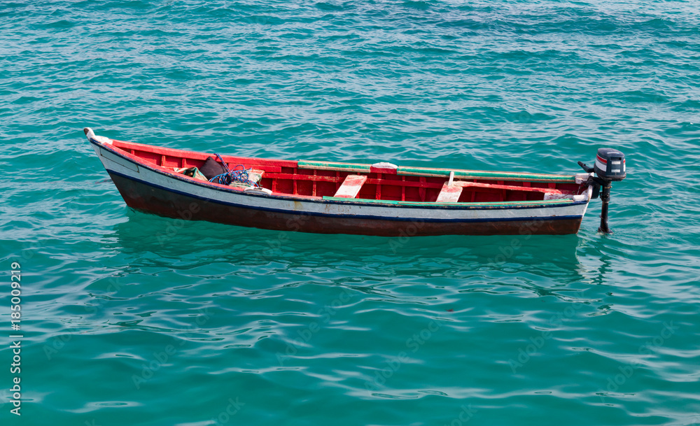 Fototapeta premium An isolated traditional red and white fishing boat floats on the clear turquoise water by the pier in Santa Maria, Sal, Cabo Verde, Africa.