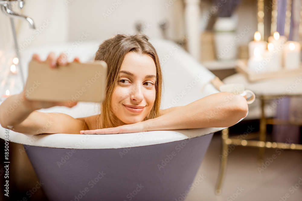 Obraz premium Young and beautiful woman making selfie photo with phone lying in the retro bath indoors