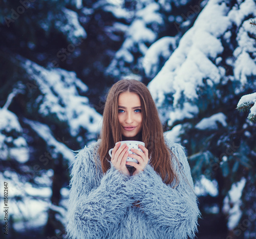 Young woman in a blue fur coat is drinking tea in the winter forest