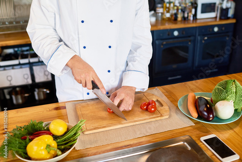 Close up of unrecognizable cook cutting fresh cherry tomatoes and other vegetables with chef knife while working in modern kitchen, copy space