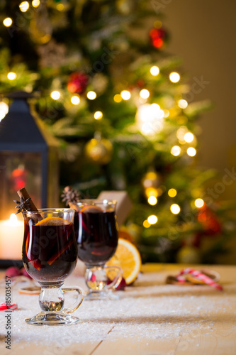 Two high glasses of christmas mulled wine with red apples, orange slices, cinnamon sticks and spices on wooden table with bokeh background. Selective focus. © Chinara