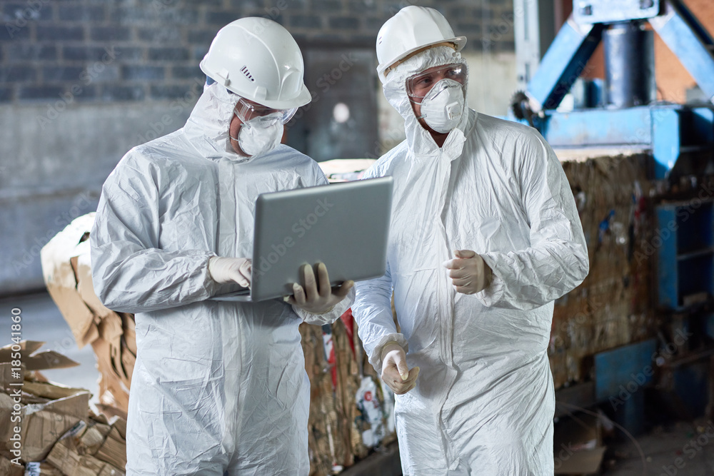 Portrait of two workers wearing biohazard suits using laptop  in industrial warehouse of modern waste processing plant against recyclable cardboard in background