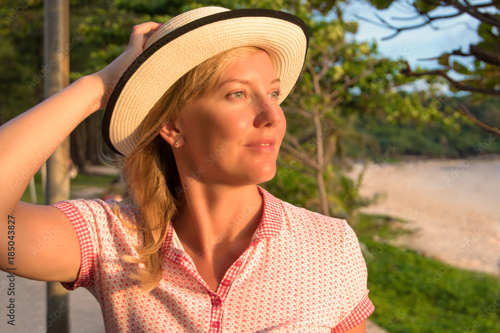 Happy woman sitting on the coast on beach looks into the distance at sea and holding her hat.